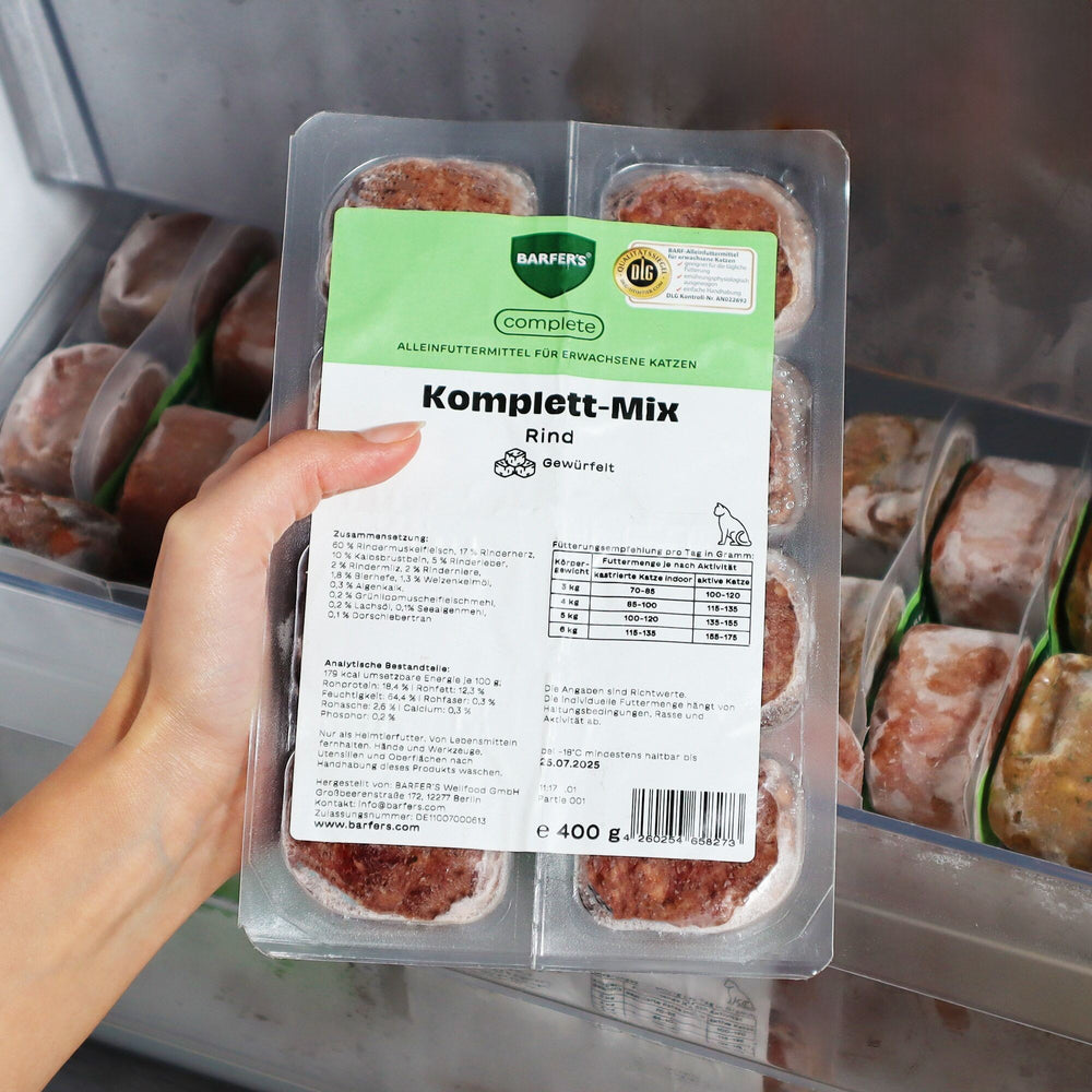 Eine Hand hält eine 400g Verpackung mit dem BARFER’S complete Komplett-Mix Rind für Katzen über ein geöffnetes Gefrierfach – sichtbar sind 8 gefrorene 50g-Würfel mit Etikett und DLG-Siegel