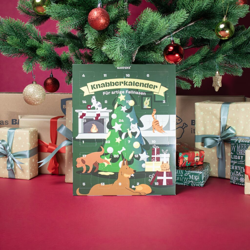Ein geschmückter Weihnachtsbaum mit Geschenken und der BARFER’S Knabberkalender für artige Fellnasen steht im Mittelpunkt, umgeben von festlich verpackten Geschenken und BARFER’S Versandkartons