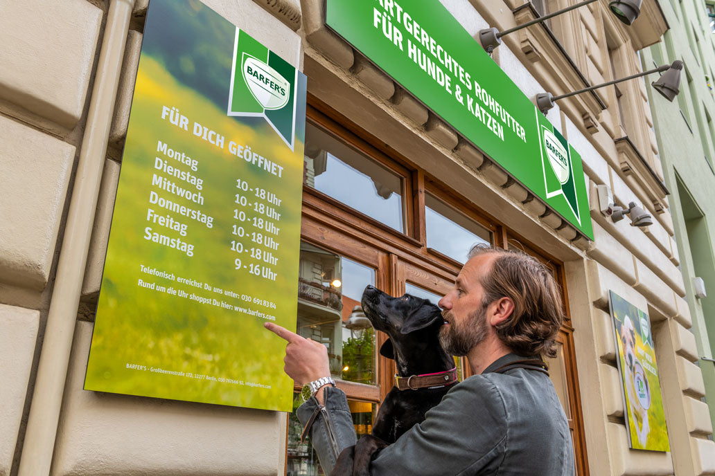 Ein weißer Mann mittleren Alters mit braunen Haaren und Bart hat einen kleinen schwarzen Hund auf dem Arm und zeigt auf das Öffnungszeiten-Schild vor dem BARFER'S Store in Berlin-Kreuzberg.