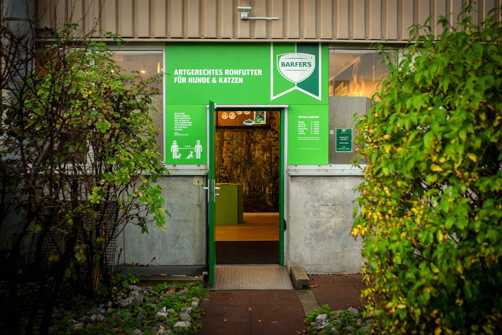 Eingang des BARFER’S Hofladens in Berlin-Marienfelde mit grünem Schild ‚Artgerechtes Rohfutter für Hunde & Katzen‘, flankiert von Büschen und offener Tür zum Innenraum
