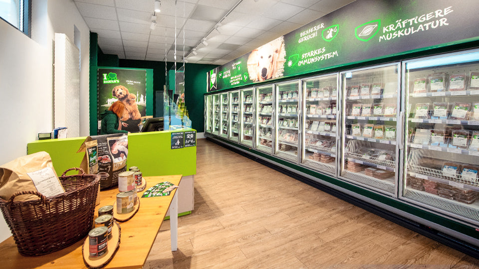 10 Tiefkühlschränke mit gefrorenem BARF-Fleisch im BARFER'S Store Berlin-Spandau. Gegenüber der Tiefkühlschränke ist der Kassenbereich, hinter dem eine rothaarige BARF-Verkäuferin steht. Daneben befindet sich ein Holztisch, auf dem BARF-Zusätze angeboten werden.