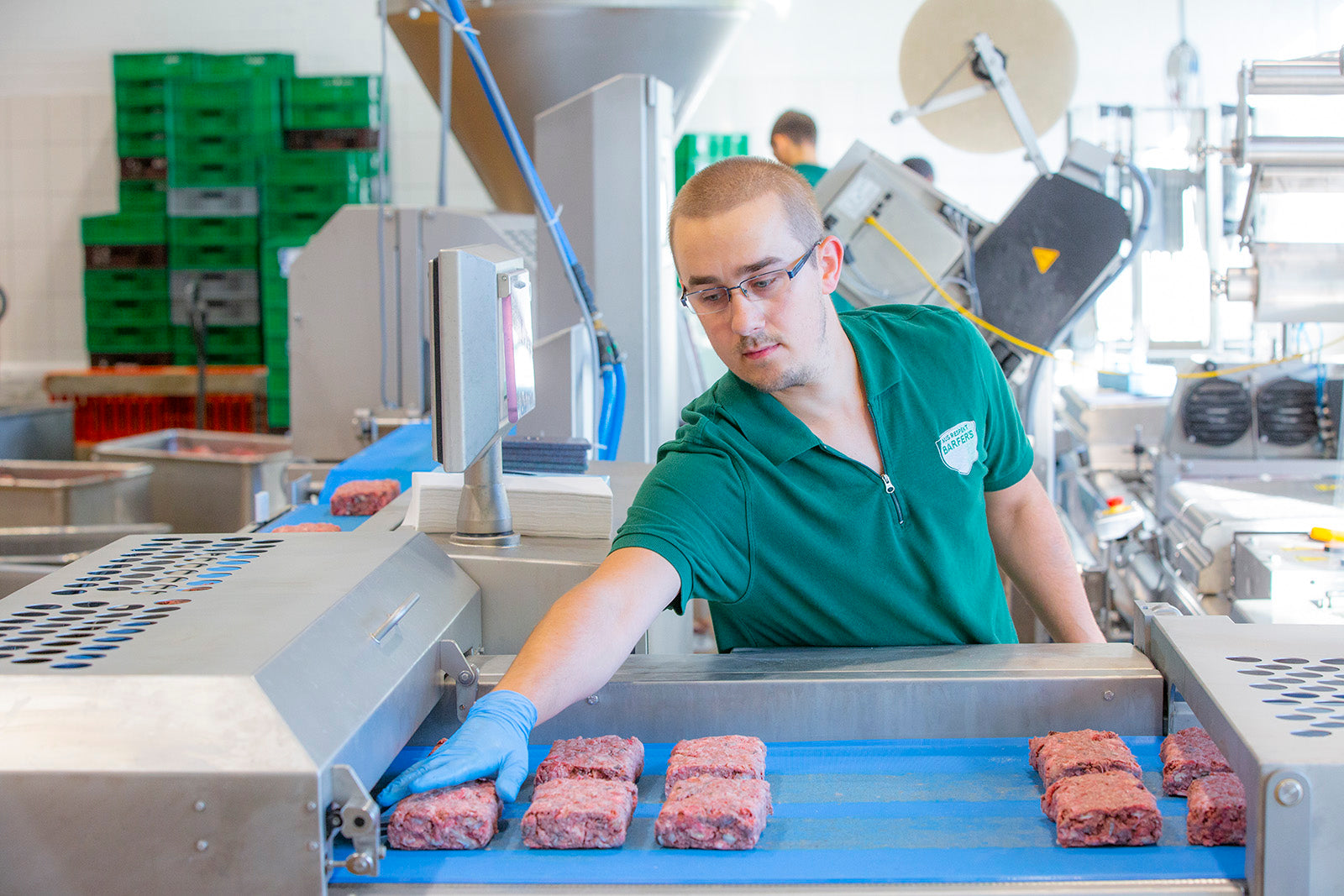 Männlicher Mitarbeiter mit blauen Einmalhandschuhen in der BARFER’S Produktion sortiert BARF-Fleischwürfel auf einem blauen Förderband, umgeben von moderner Verarbeitungstechnik und grünen Transportkisten im Hintergrund