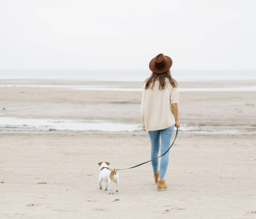 Frau mit braunem Hut spaziert entspannt mit ihrem kleinen Hund an der Leine am Strand, im Hintergrund das ruhige Meer