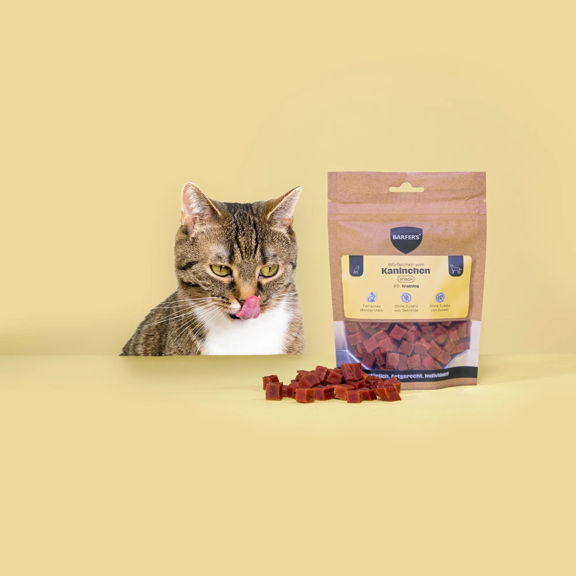 Eine grau-schwarz gestreifte Katze mit weißer Brust leckt sich das Maul und schaut Leckerli vom Kaninchen der Marke BARFER'S an, die neben einer vollen Verpackung "Würfelchen vom Kaninchen" liegen