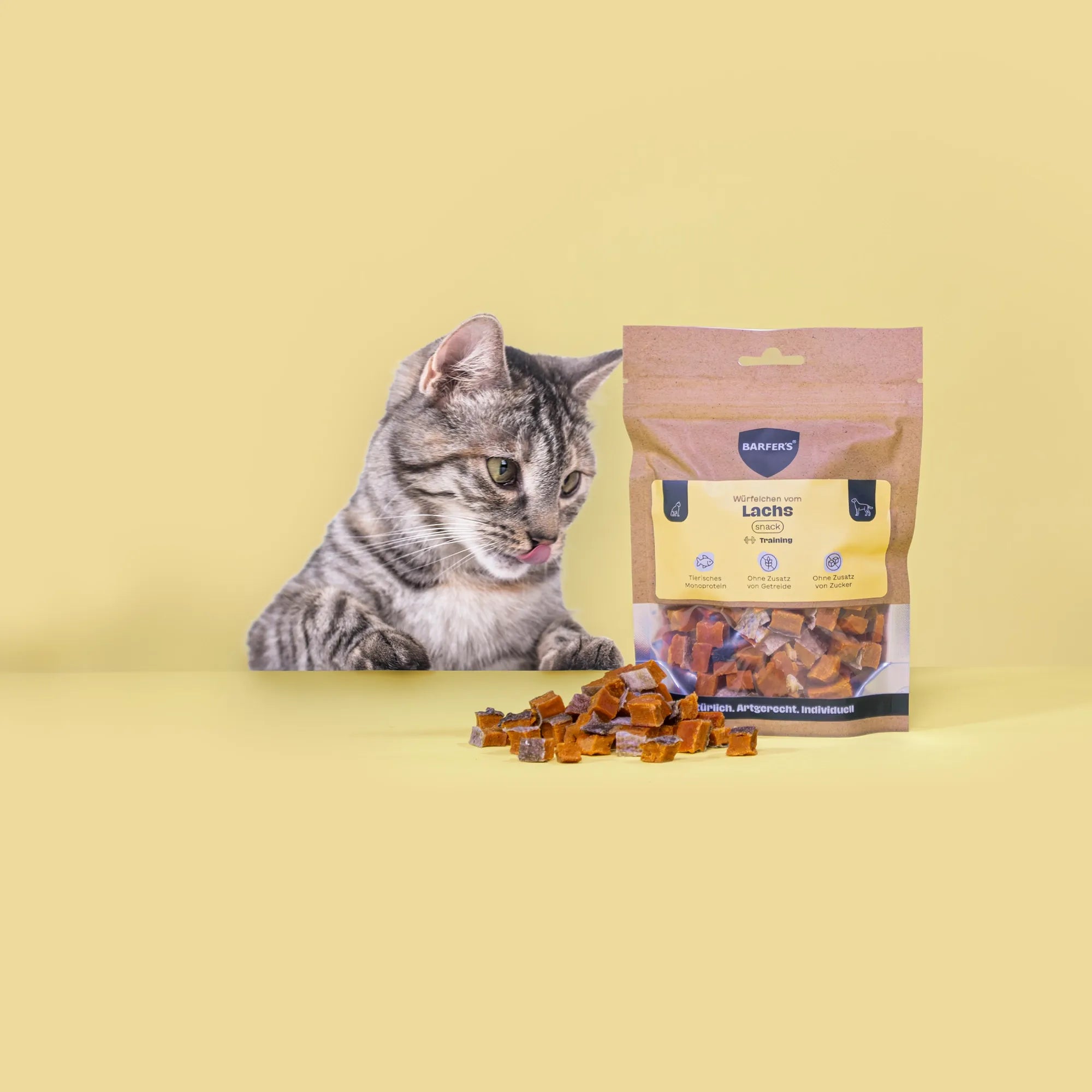 Eine schwarz-grau gestreifte Katze leckt sich das Maul und schaut Leckerli vom Lachs der Marke BARFER'S an, die neben einer vollen Verpackung "Würfelchen vom Lachs" liegen
