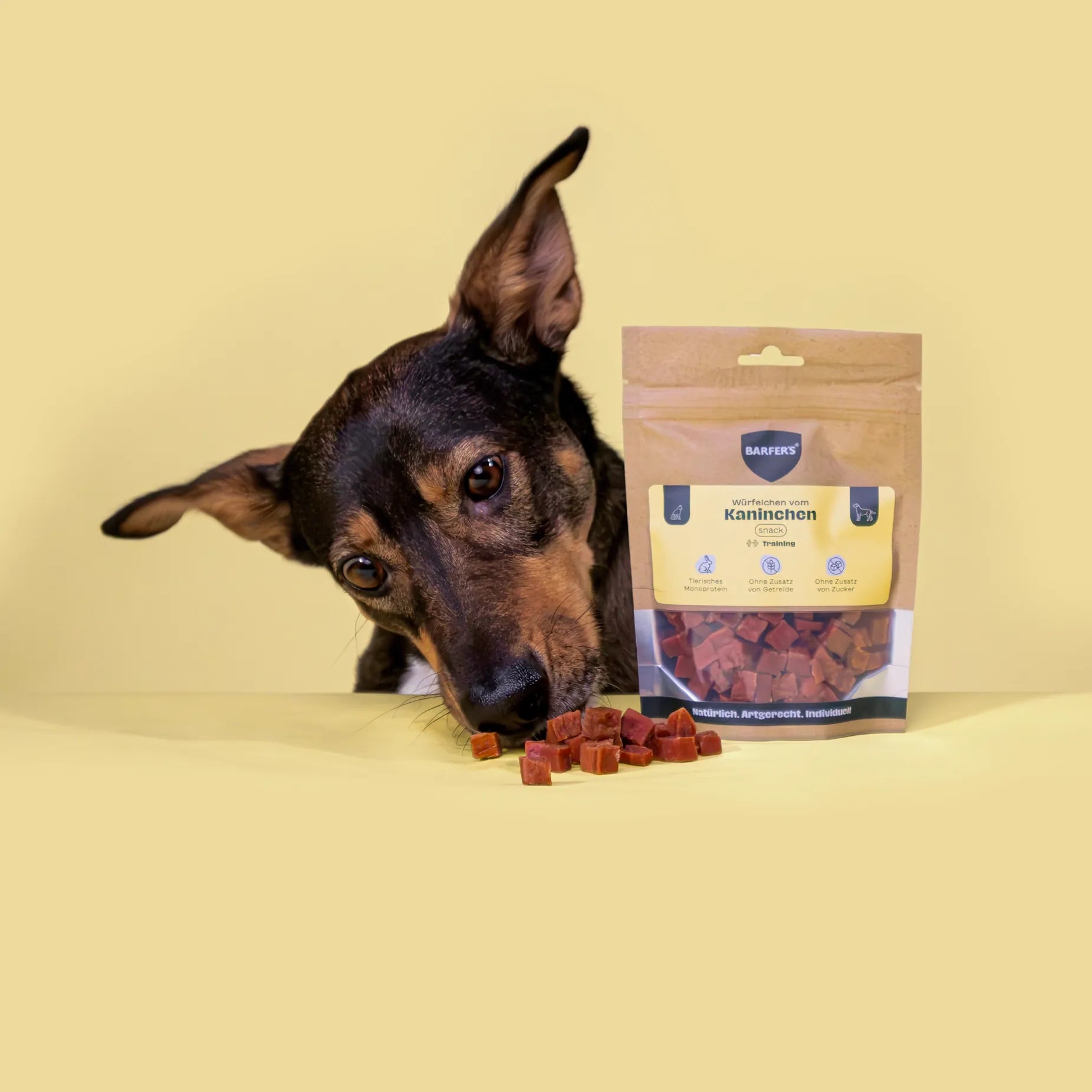 Ein schwarz-brauner Hund beschnuppert Leckerli vom Kaninchen der Marke BARFER'S, die neben einer vollen Verpackung "Würfelchen vom Kaninchen" liegen