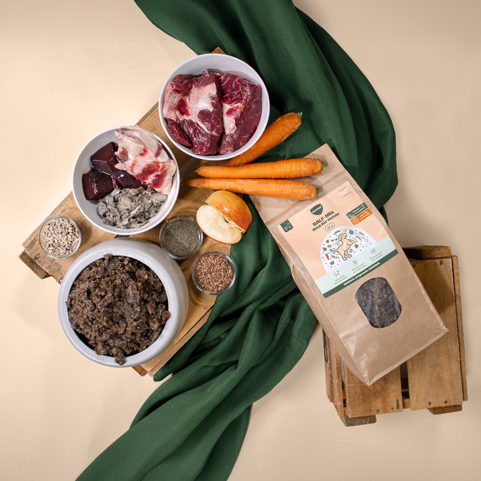Zutaten des BARFER’S BALF-Mix Rind für Hunde – Rindermuskelfleisch, Rinderpansen, Rinderblättermagen, Rinderlunge, Rinderleber, Rindermilz, Rinderniere, Rinderknochen sowie pflanzliche Komponenten wie Leinkuchen, Algenkalk, Sanddorntrester, Apfeltrester, Möhrentrester und Aroniatrester, angerichtet mit Möhren, Apfel, Sonnenblumenkernen und verschiedenen Saaten neben einer Packung des luftgetrockneten BARF-Mixes