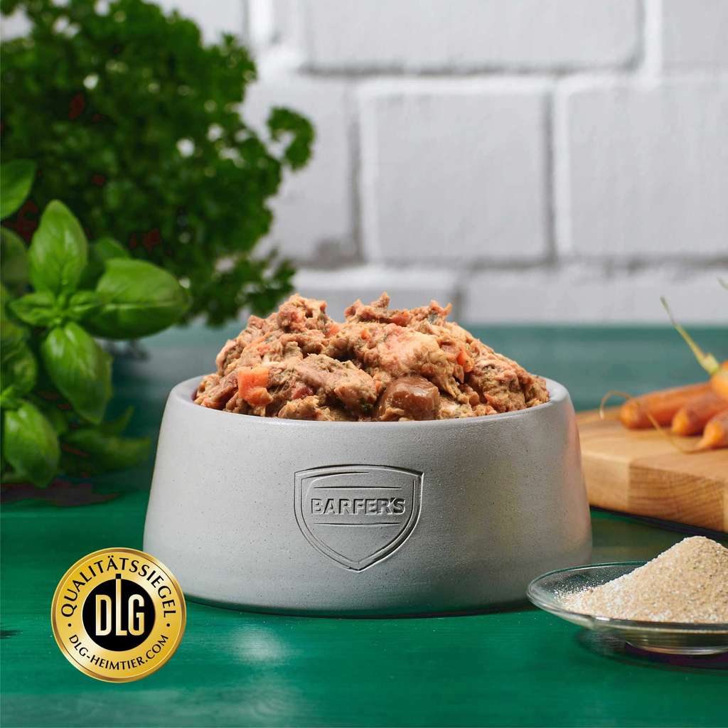 Gefüllter Napf mit BARFER’S Komplett-Mix Junior – erkennbar frisches Gemüse und Fleischstücke, daneben DLG-Siegel, im Hintergrund Gemüse und Kräuter