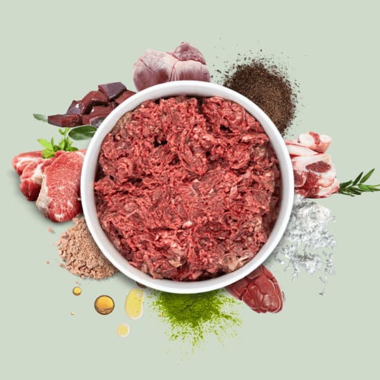 BARFER’S Komplett-Mix im Napf, umgeben von frischen Zutaten wie Muskelfleisch, Innereien, Knochen, Kräutern, Ölen und Mineralien – vollwertiges Fertigbarf für Katzen