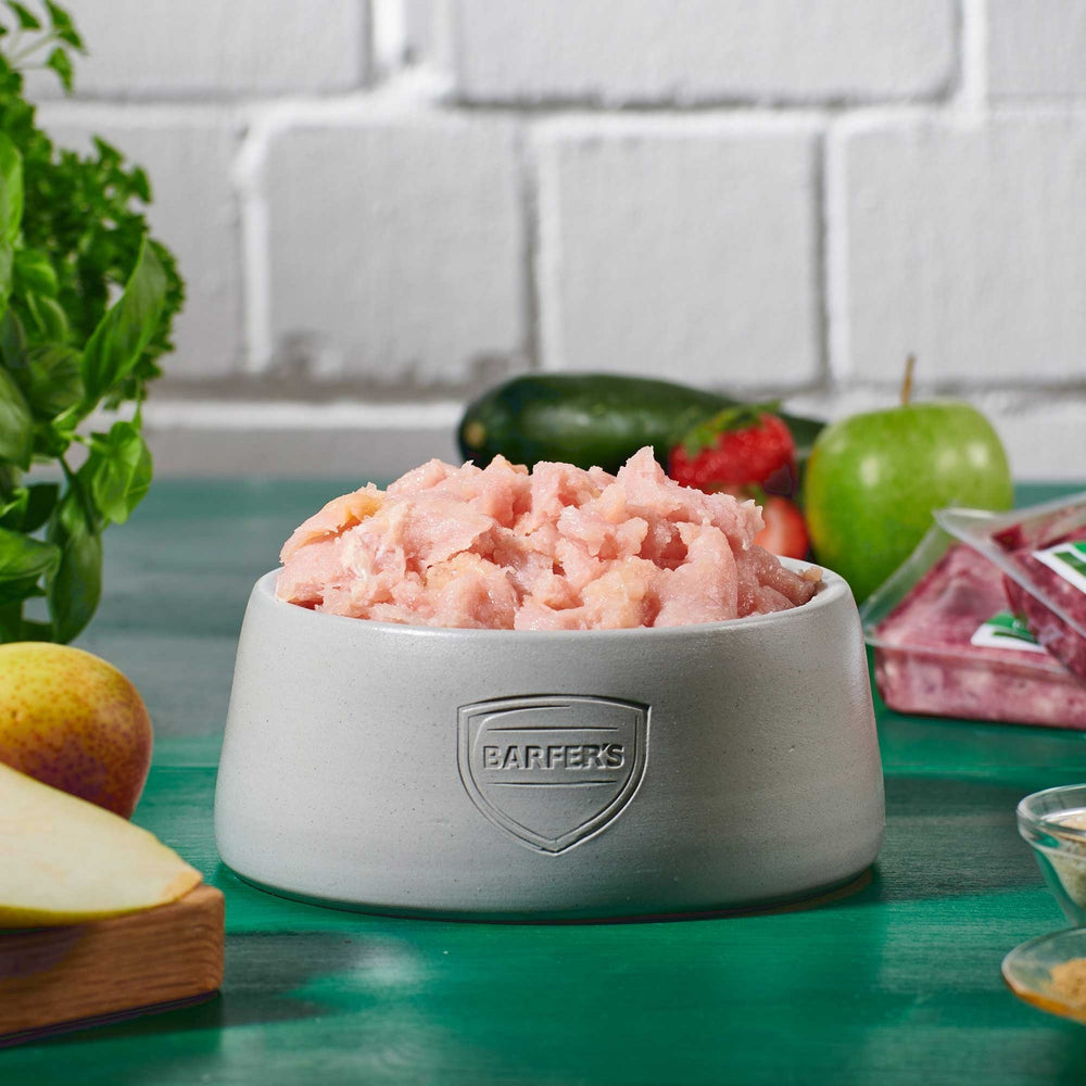 Grauer Futternapf mit BARFER’S Logo, gefüllt mit gewolftem BARF-Truthahn Muskelfleisch, im Hintergrund frisches Obst und Gemüse auf grünem Holztisch