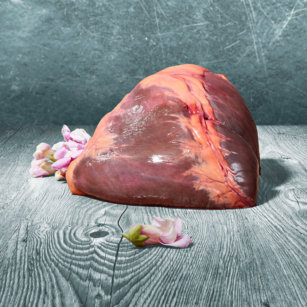 Ganzes BARF-Pferdeherz auf grauer Holzoberfläche, dekoriert mit zartrosa Blüten, vor grauem Hintergrund