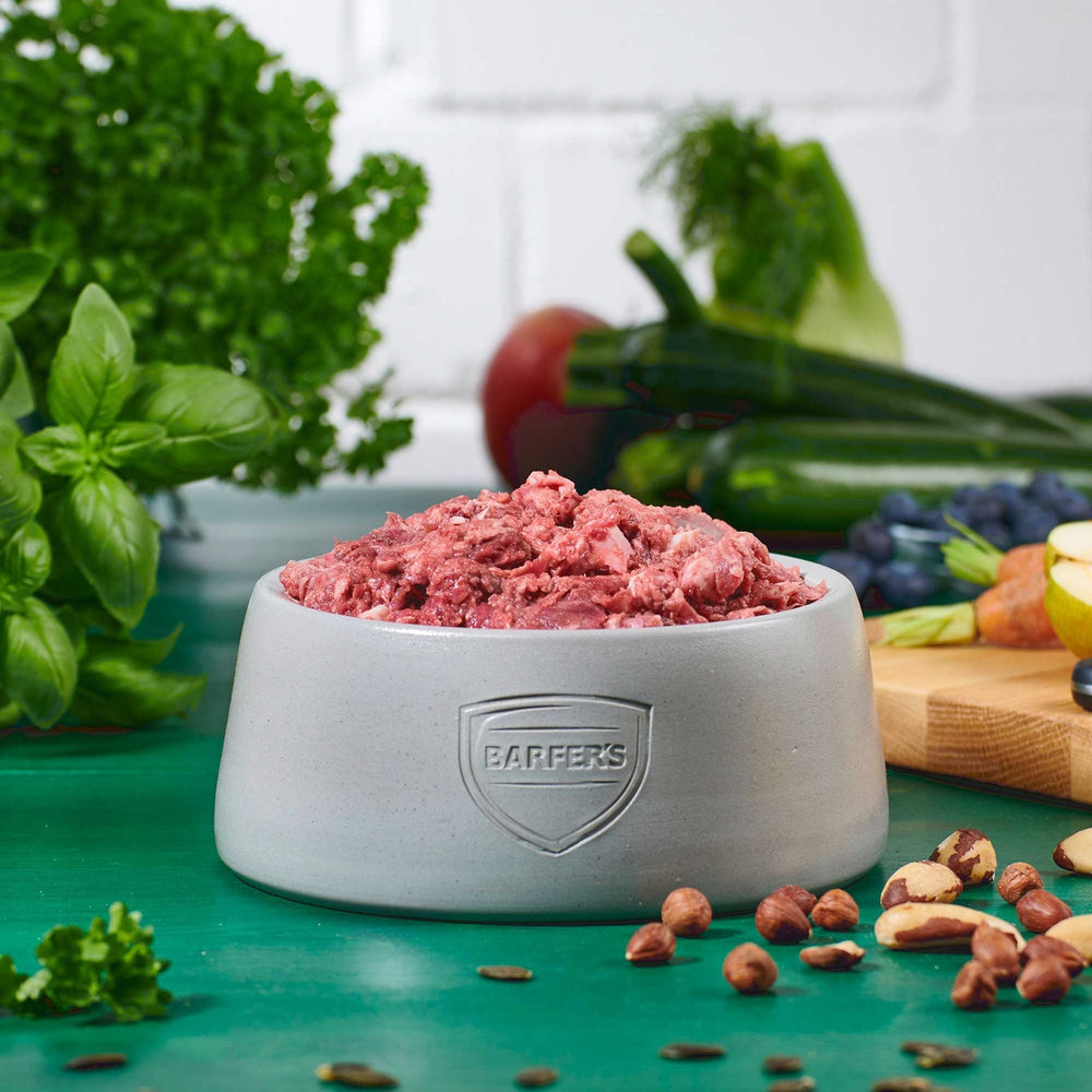 Hellgrauer Keramiknapf mit BARFER’S Logo, gefüllt mit Fleisch-Mix Lamm mit Rind & Huhn– aus Lammfleisch, Rinderpansen, Hühnerhals, Lammlunge, Lammherz und Lammleber, dekoriert mit frischem Gemüse, Apfelhälfte, Blaubeeren, Möhren, Kräutern und Nüssen