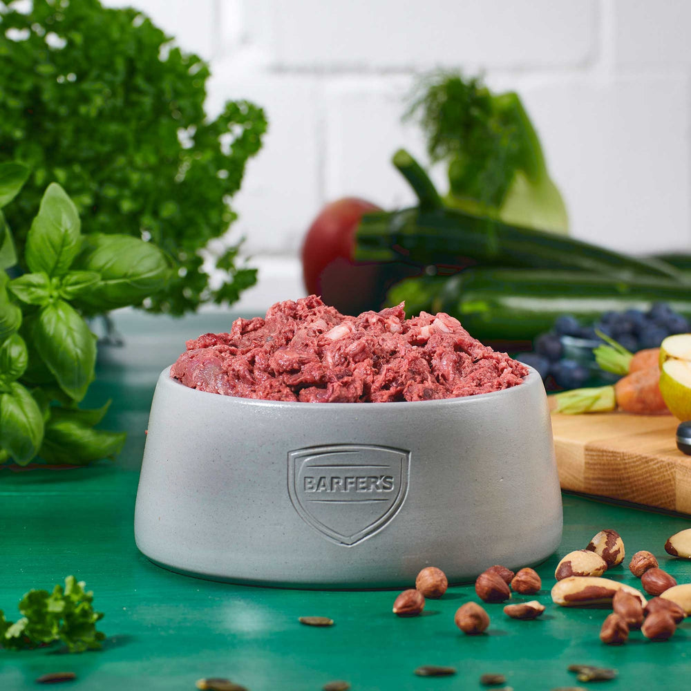 Hellgrauer Keramiknapf mit BARFER’S Logo, gefüllt mit Fleisch-Mix Wild, Lamm, Rind & Geflügel – aus Hirschfleisch, Lammpansen, Hühnerhals, Rinderlunge, Lammleber und Putenherz, dekoriert mit frischem Gemüse, Apfelhälfte, Blaubeeren, Möhren, Kräutern und Nüssen