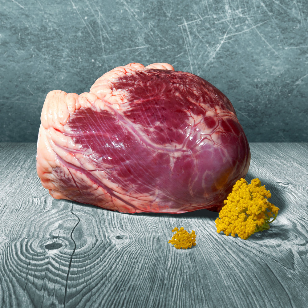 Ganzes rohes BARF-Rinderherz mit sichtbarer Fettschicht auf Holztisch, dekoriert mit gelben Blüten