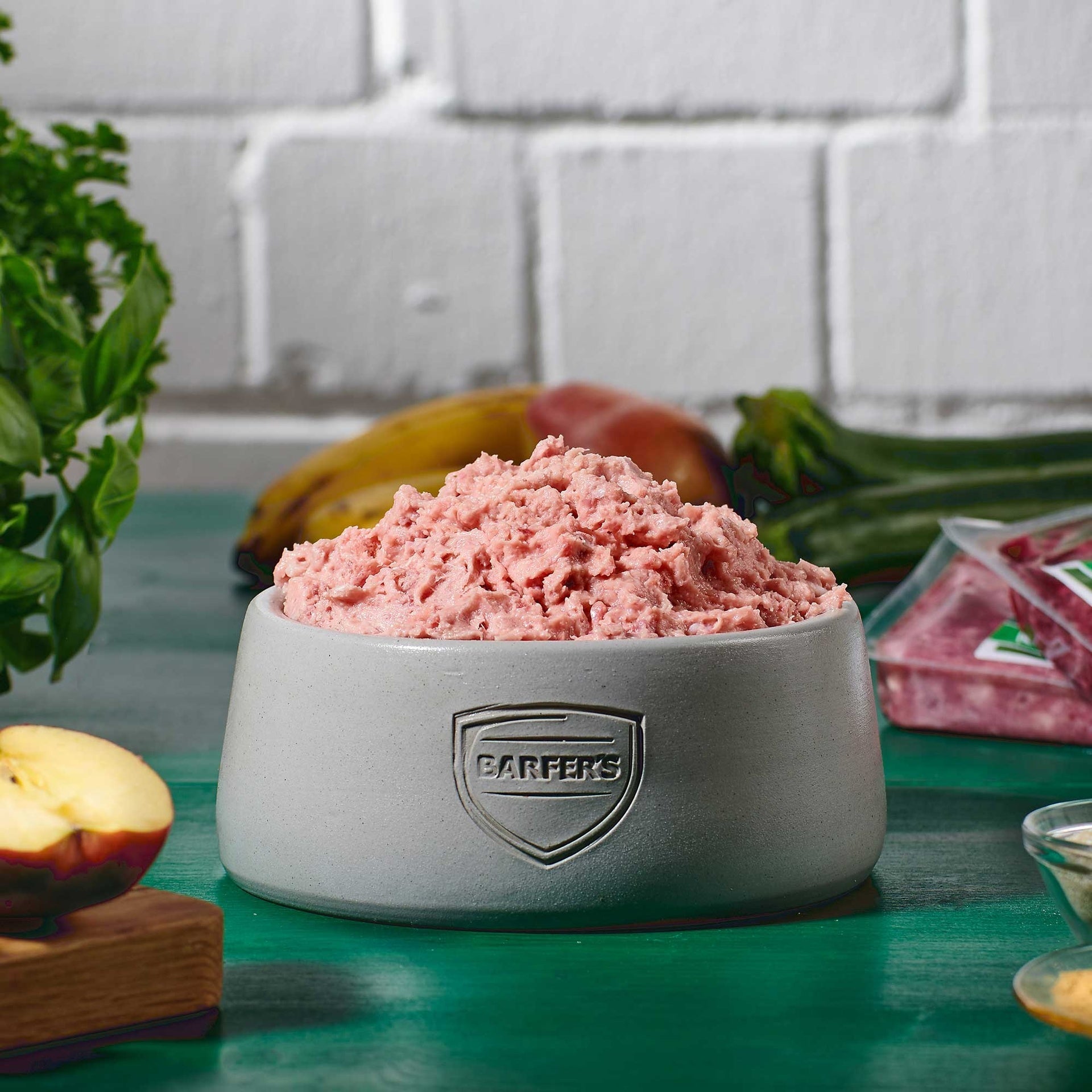 Grauer Futternapf mit BARFER’S Logo, gefüllt mit gewolften Hühnerhälsen, im Hintergrund Obst und Gemüse auf grünem Holztisch