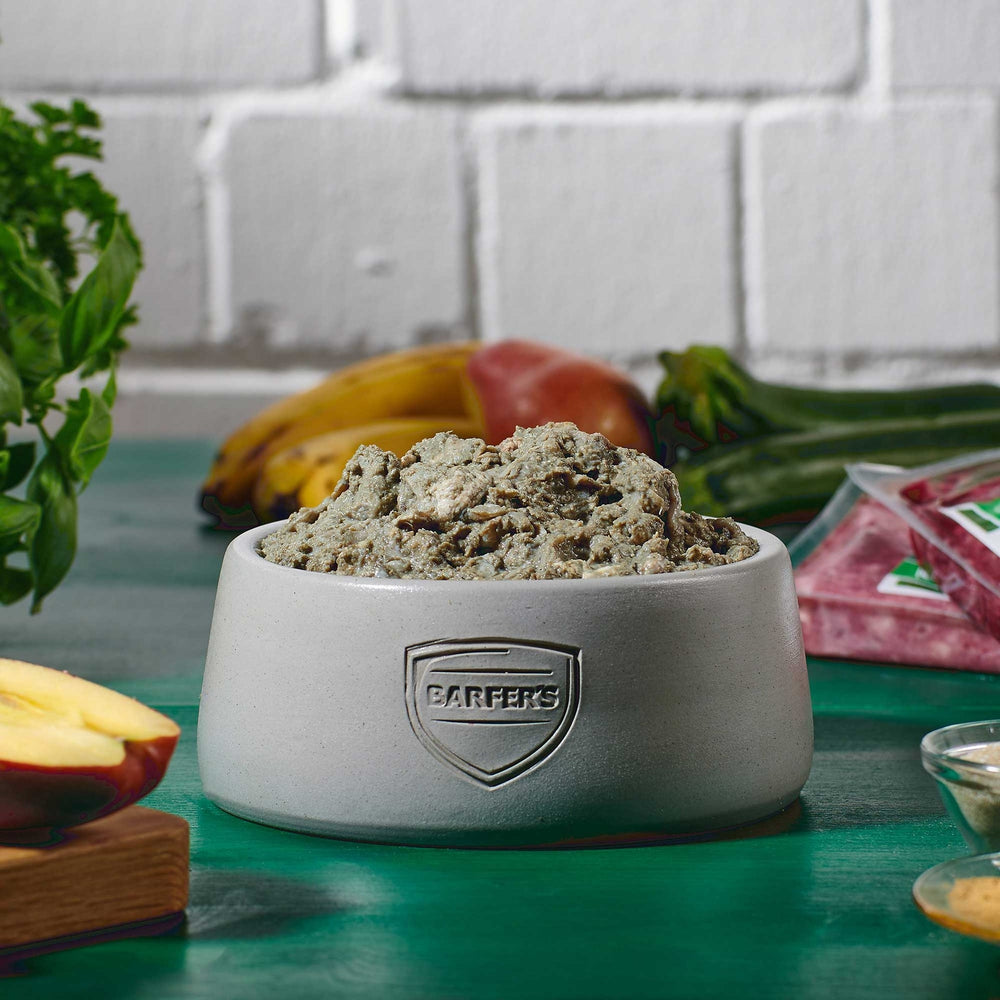 Grauer Futternapf mit BARFER’S Logo, gefüllt mit gewolftem Lammpansen, daneben Obst und Gemüse auf grünem Tisch