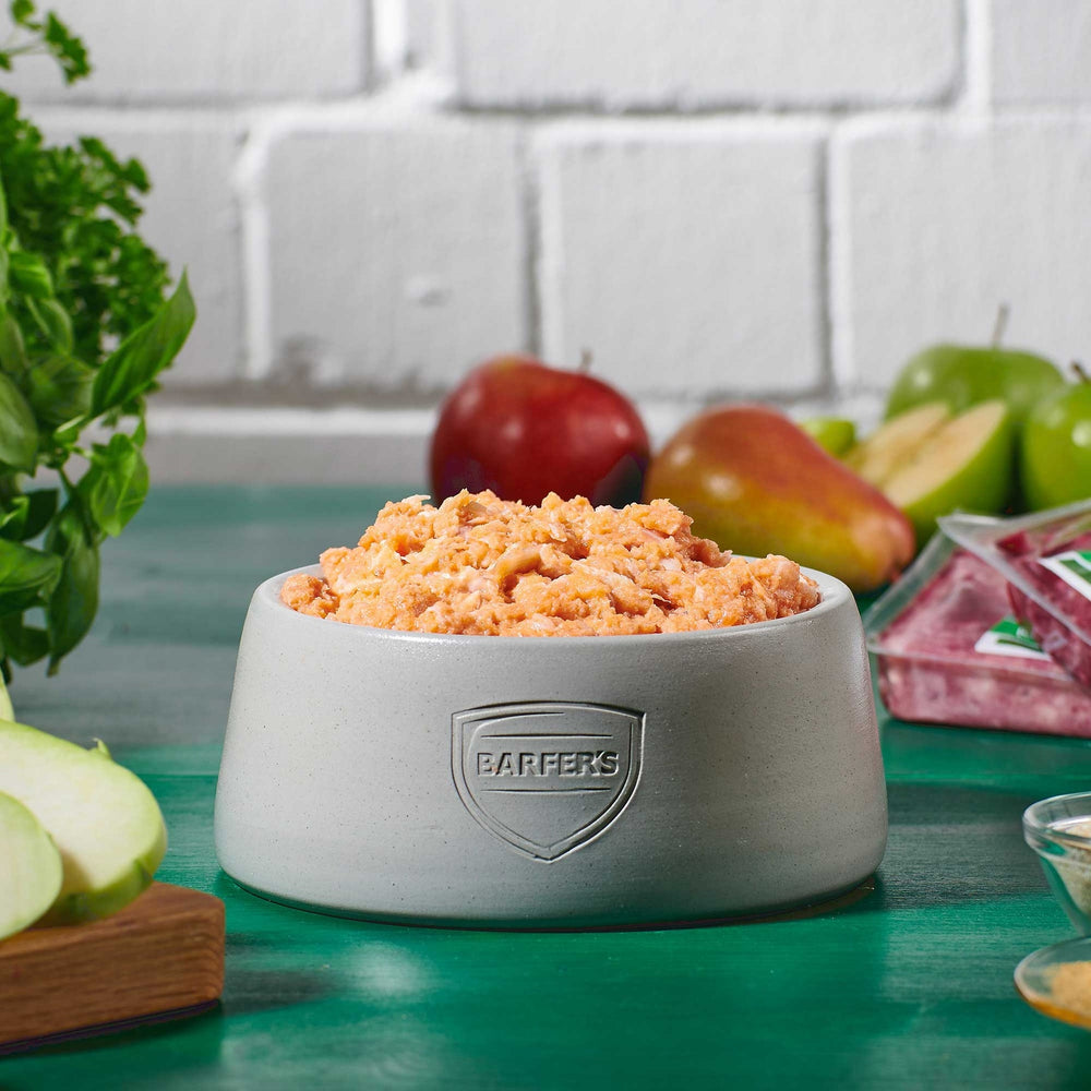Grauer Futternapf mit BARFER’S Logo, gefüllt mit gewolftem BARF-Lachs, umgeben von Äpfeln, Birnen und frischen Kräutern auf grünem Holztisch