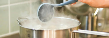 Hand hält einen Schöpflöffel über einem dampfenden Kochtopf – Symbolbild fürs Kochen von BARF-Fleisch