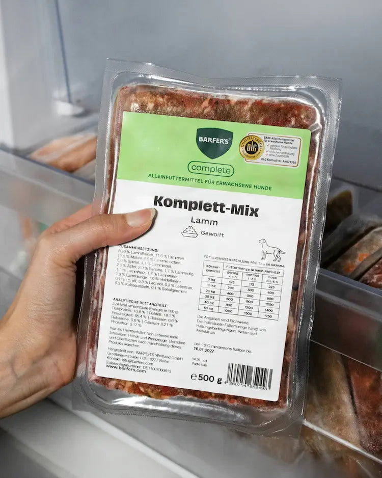 Hand hält eine 500-g-Packung BARFER’S complete Komplett-Mix Lamm, gewolft, mit DLG-Siegel vor einem geöffneten Gefrierfach, DLG-zertifiziertes FertigBARF als Alleinfuttermittel für erwachsene Hunde