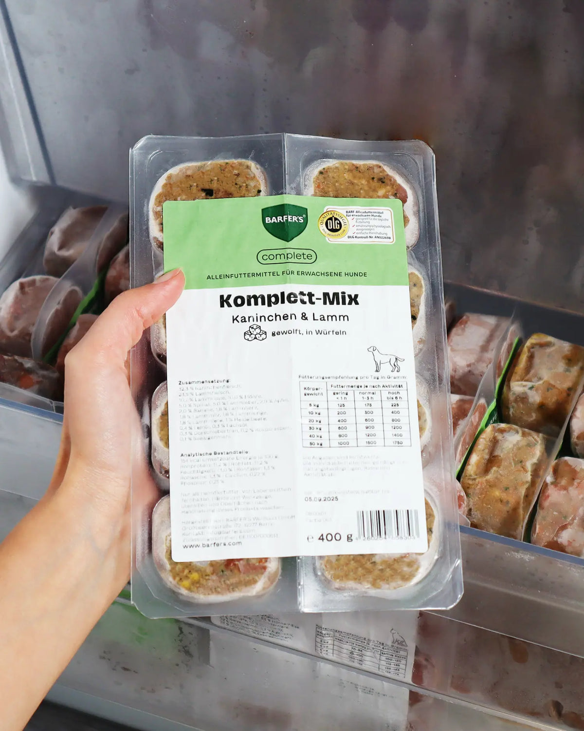 Hand hält BARFER'S Komplett-Mix Kaninchen & Lamm, DLG-zertifiziertes Fertigbarf mit Fütterungstabelle, vor geöffnetem Gefrierfach mit weiteren Würfel-Portionen