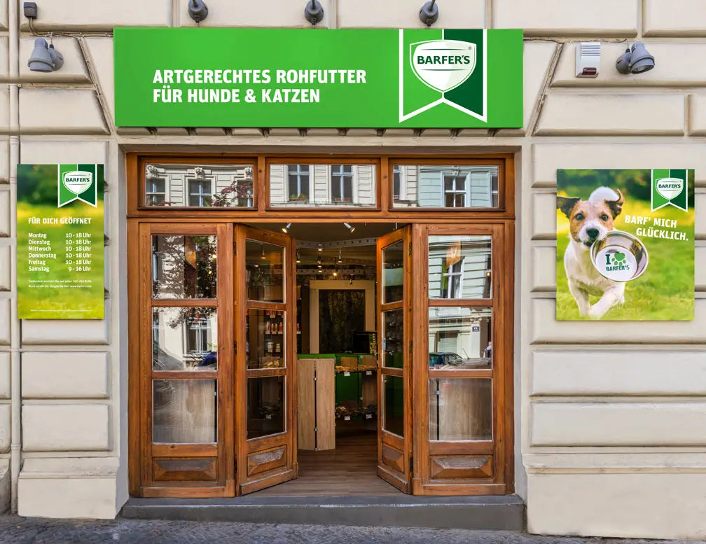Außenansicht eines BARFER’S Stores in einem hellen Altbau mit geöffneter, holzgerahmter Eingangstür. Über dem Eingang befindet sich ein grünes Schild mit dem Schriftzug „Artgerechtes Rohfutter für Hunde & Katzen“ und dem BARFER’S Logo. Links hängt ein Schild mit den Öffnungszeiten, rechts ein Werbeplakat mit einem Hund und einer Futterschüssel.