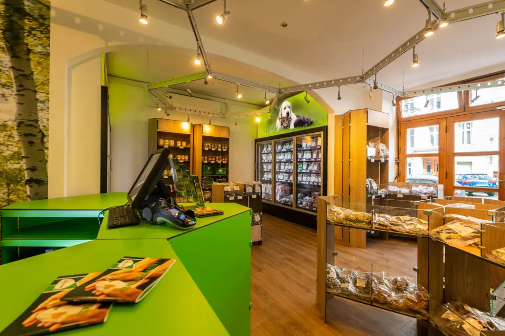 Innenansicht eines BARFER’S Stores mit grüner Verkaufstheke und Kasse im Vordergrund. Im Verkaufsraum stehen Holzregale mit Tierfutterdosen und weitere Regale mit verpackten Produkten. Eine Kühltheke mit Glastüren befindet sich an der Rückwand, darüber ist ein Bild mit einem Hund zu sehen. Rechts stehen mehrere Verkaufsvitrinen mit verpackten Kauartikeln und Snacks für Hunde. Durch die großen Fenster fällt Tageslicht in den Raum.