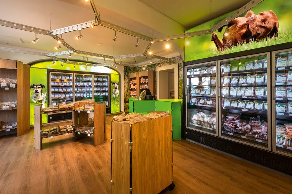 Innenansicht des BARFER’S Stores in Berlin Kreuzberg mit Kühlregalen voller Frischfleisch-Portionen, Holzregalen mit Snacks und einem Wandbild mit Hunden auf grüner Wiese.