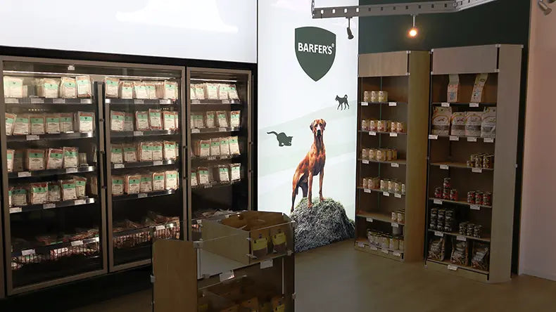 Innenansicht des BARFER'S Hofladens Berlin-Marienfelde: Tiefkühlregale mit BARF-Produkten, daneben Trocken-BARF und Leckerli-Regale sowie ein Wandbild mit BARFER'S Logo und Hund.