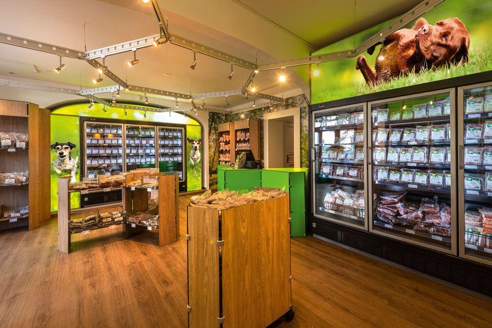 Holz-Leckerli-Regale in der Mitte der Verkaufsfläche im BARFER'S Store Berlin-Kreuzberg. Links und rechts außen befinden sich beleuchtete Tiefkühlregale mit BARF-Produkten für Hunde und Katzen.