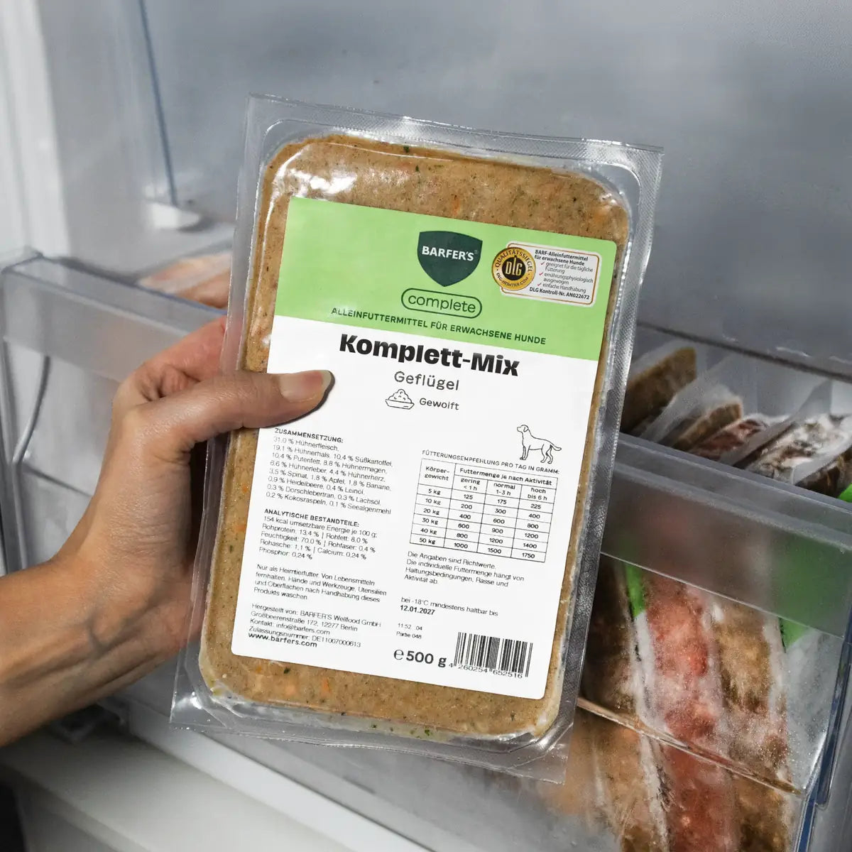 Eine Hand hält eine 500g-Packung BARFER’S Komplett-Mix Geflügel für Hunde vor ein geöffnetes Gefrierfach, sichtbar sind das grüne complete-Label, das DLG-Qualitätssiegel und weitere gefrorene BARF-Packungen im Hintergrund
