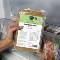 Eine Hand hält eine 500g-Packung BARFER’S Komplett-Mix Geflügel für Hunde vor ein geöffnetes Gefrierfach, sichtbar sind das grüne complete-Label, das DLG-Qualitätssiegel und weitere gefrorene BARF-Packungen im Hintergrund