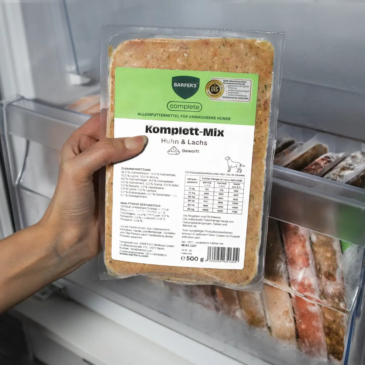 Eine Hand hält eine 500g-Packung BARFER’S Komplett-Mix Huhn & Lachs für Hunde im Gefrierfach, mit grünem complete-Label und sichtbarem DLG-Qualitätssiegel