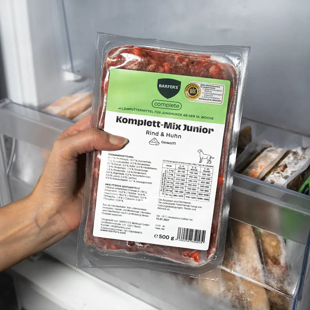 Eine Hand präsentiert eine 500g-Packung BARFER’S Komplett-Mix Junior Rind & Huhn für Junghunde im Gefrierfach, mit DLG-Siegel und Hinweis auf Alleinfuttermittel ab der 14. Woche