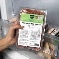 Eine Hand präsentiert eine 500g-Packung BARFER’S Komplett-Mix Junior Rind & Huhn für Junghunde im Gefrierfach, mit DLG-Siegel und Hinweis auf Alleinfuttermittel ab der 14. Woche