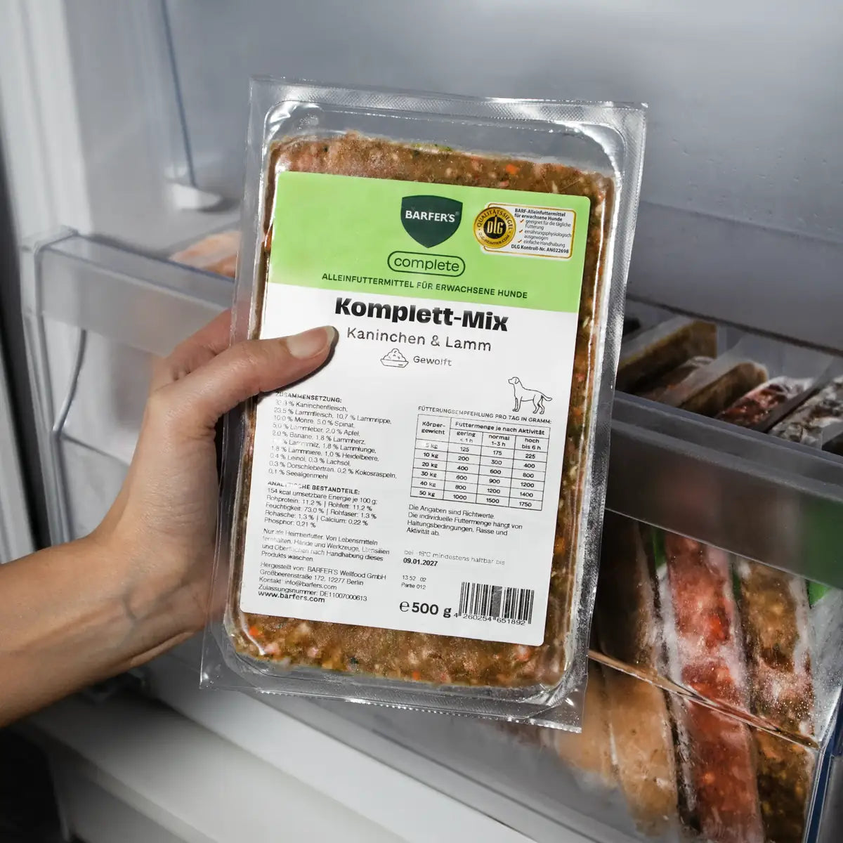 Eine Hand hält eine 500g-Packung BARFER’S Komplett-Mix Kaninchen & Lamm für Hunde im Gefrierfach, mit grünem Label, DLG-Zertifizierung und gewolftem Fertigbarf