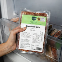 Eine Hand hält eine 500g-Packung BARFER’S Komplett-Mix Lamm für Hunde im Gefrierfach, sichtbar sind das complete-Label und das DLG-Qualitätssiegel