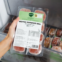 Eine Hand hält eine 400 g Verpackung mit dem BARFER’S complete Würfel Komplett-Mix Rind & Geflügel für Katzen über ein geöffnetes Gefrierfach – sichtbar sind 8 gefrorene 50 g-Würfel mit Etikett und DLG-Siegel