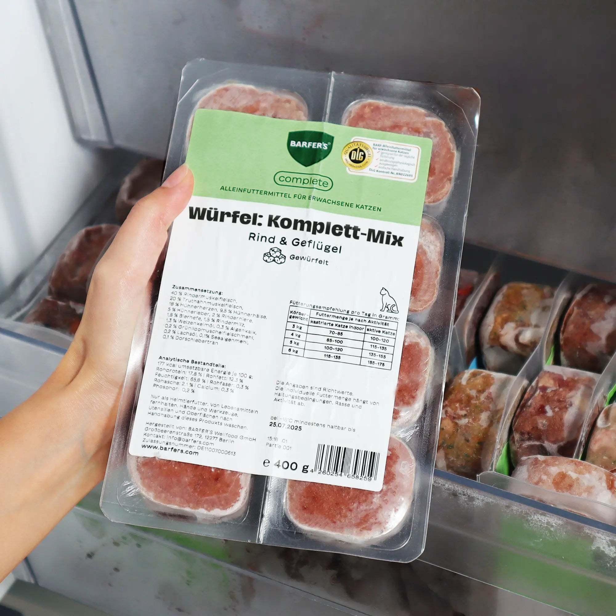 Eine Hand hält eine 400 g Verpackung mit dem BARFER’S complete Würfel Komplett-Mix Rind & Geflügel für Katzen über ein geöffnetes Gefrierfach – sichtbar sind 8 gefrorene 50 g-Würfel mit Etikett und DLG-Siegel