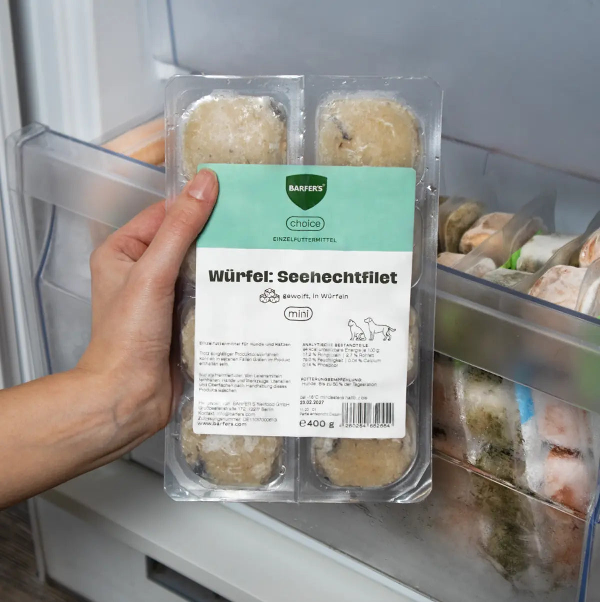 Eine Hand hält eine 400g Verpackung mit BARFER’S Choice Würfel Seehechtfilet vor ein Gefrierfach – sichtbar sind acht gefrorene Fischwürfel in transparenter Verpackung mit grün-weißem Etikett, geeignet für Hunde und Katzen
