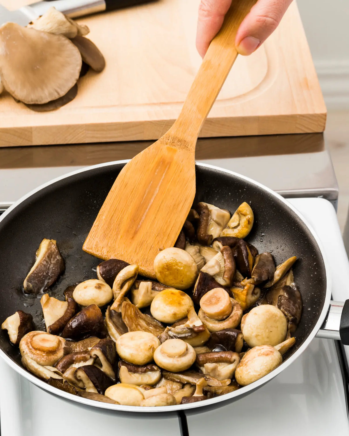 Hand rührt mit einem Holzspatel gebratene braune und weiße Champignons in einer Pfanne, im Hintergrund weitere frische Pilze auf einem Schneidebrett