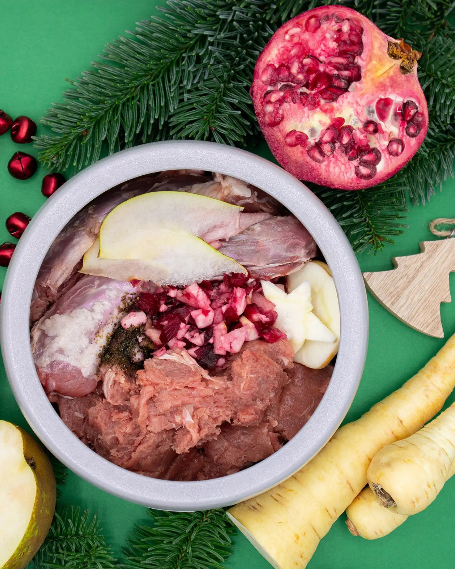 Napf mit BARF-Portion aus Fleisch, Knochen, Apfel und Granatapfelkernen neben einem aufgeschnittenen Granatapfel und Pastinaken, dekoriert mit Tannenzweigen