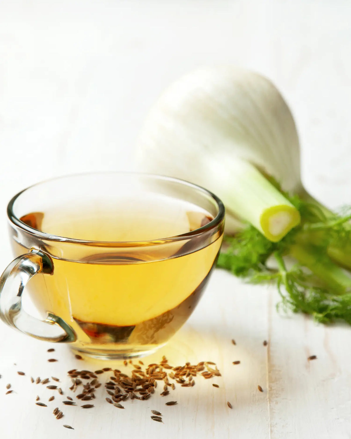 Glastasse mit Fencheltee neben einer angeschnittenen Fenchelknolle und verstreuten Fenchelsamen