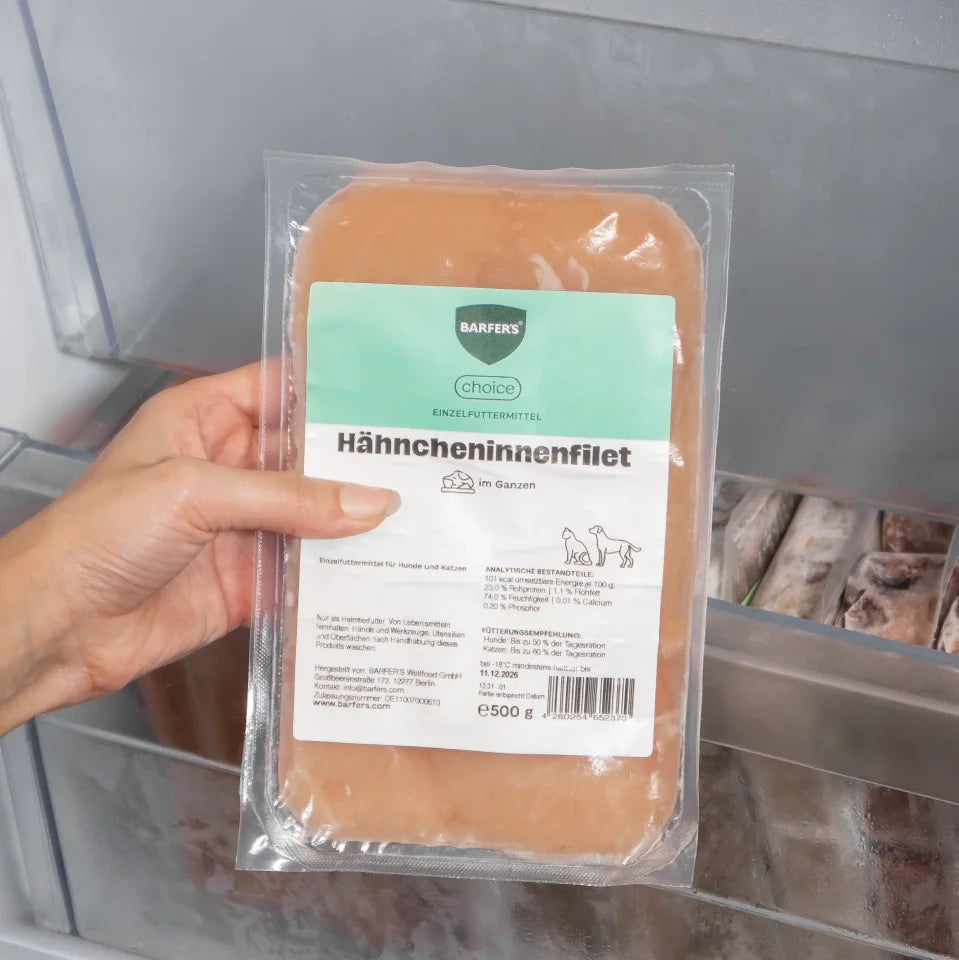 Eine Hand hält eine 500 g Verpackung BARFER’S Choice Hähncheninnenfilet im Ganzen vor ein geöffnetes Gefrierfach – Einzelfuttermittel für Hunde und Katzen