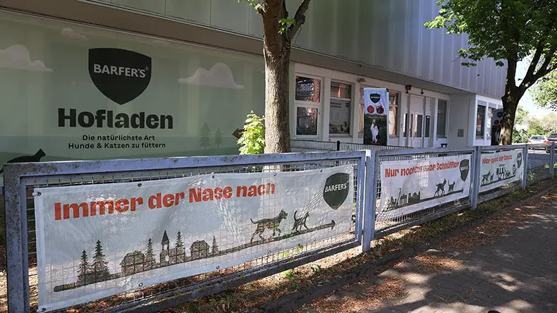 Außenansicht des BARFER'S Hofladens in Berlin-Marienfelde mit Fassadenschriftzug „Hofladen – Die natürlichste Art Hunde & Katzen zu füttern" und Werbebannern am Zaun mit der Aufschrift „Immer der Nase nach" und „Nur noch schnüffeln".