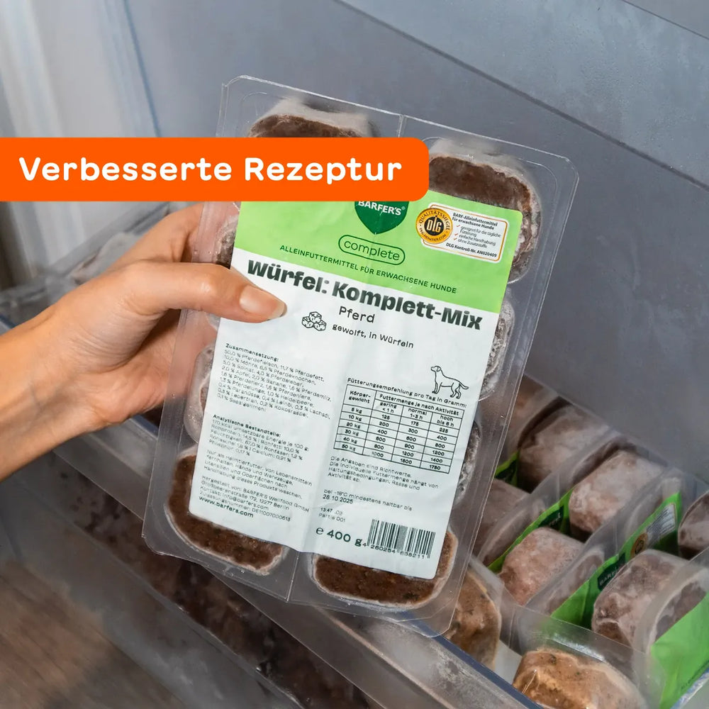 Eine Hand hält eine 400g Verpackung vom BARFER’S complete Würfel Komplett-Mix Pferd für erwachsene Hunde vor ein geöffnetes Gefrierfach – sichtbar sind 8 gefrorene 50g-Würfel mit Etikett, DLG-Siegel und dem Hinweis „Verbesserte Rezeptur“