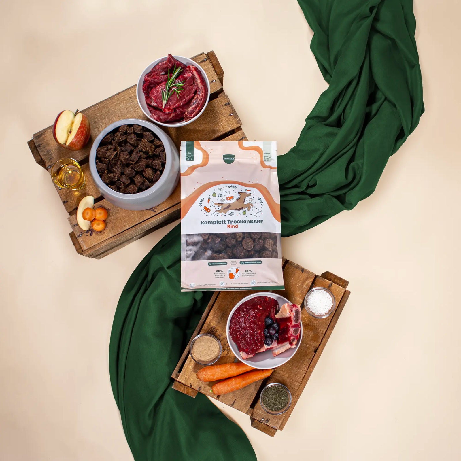 BARFER’S Komplett-TrockenBARF Rind Verpackung umgeben von frischen BARF-Zutaten wie rohem Rindfleisch, Innereien, Knochen, Karotten, Apfel, Beeren, Öl und Kräutern auf Holzbrettern mit grünem Tuch
