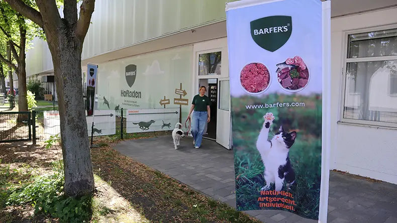 Kundin mit Hund verlässt den BARFER'S Hofladen in Berlin-Marienfelde – im Hintergrund die Außenfassade mit Hofladen-Schild, im Vordergrund ein Fahne mit BARF-Produkten und einer Katze.