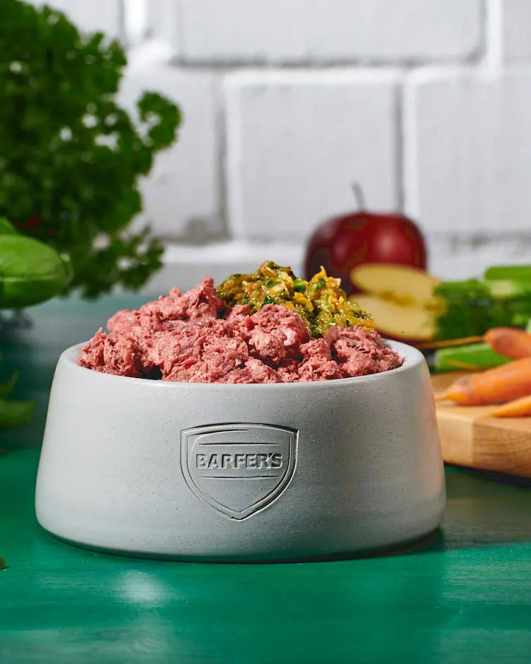 Grauer BARFER’S Keramiknapf mit gewolftem rohem Fleisch und püriertem Gemüse, im Hintergrund frische Möhren, ein Apfel und Kräuter auf einem grünen Tisch