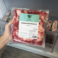 Eine Hand hält eine 1000g Verpackung BARFER’S Choice Rinderstichfleisch im Stück für Hunde und Katzen vor einem geöffneten Gefrierfach