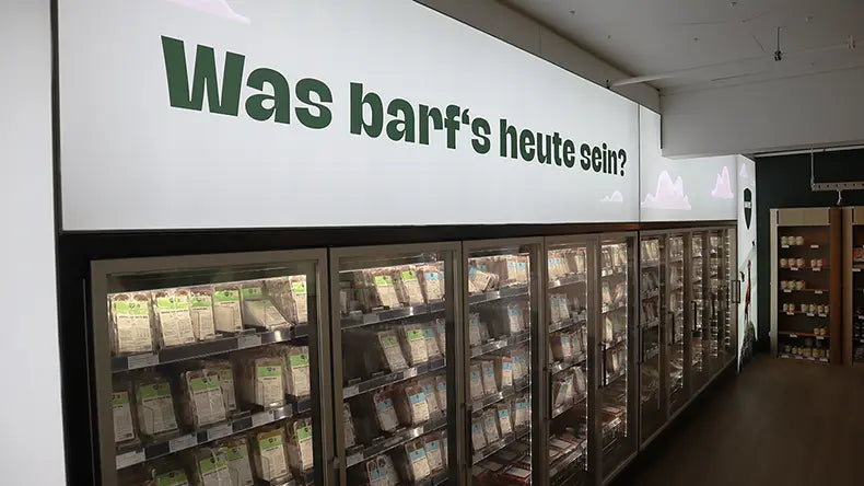 Tiefkühlregal im BARFER'S Hofladen Berlin-Marienfelde mit gefüllten Kühlschränken und dem Wandschriftzug „Was barf's heute sein?"
